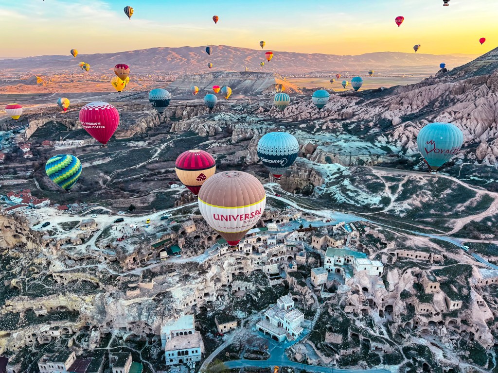 Birds eye view of dozens of hot air balloons in Cappadocia, Turkey over a nearby cave village at sunrise.